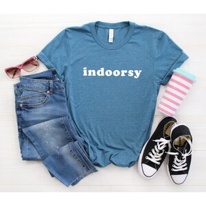 K&ouml;nnte beinhalten: Ein blaues T-Shirt mit dem Wort "indoorsy" in wei&szlig; auf der Vorderseite. Das Shirt liegt flach auf einer wei&szlig;en Oberfl&auml;che mit einer blauen Jeans, einer Sonnenbrille und einer rosa-wei&szlig; gestreiften Tasse.