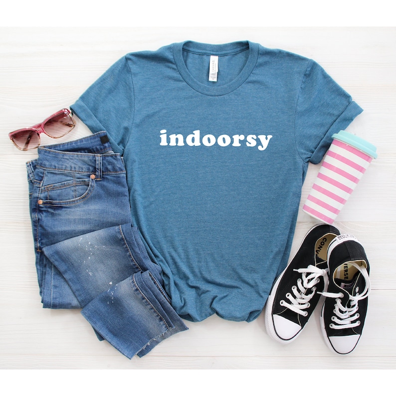 K&ouml;nnte beinhalten: Ein blaues T-Shirt mit dem Wort "indoorsy" in wei&szlig; auf der Vorderseite. Das Shirt liegt flach auf einer wei&szlig;en Oberfl&auml;che mit einer blauen Jeans, einer Sonnenbrille und einer rosa-wei&szlig; gestreiften Tasse.