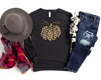 May include: A dark grey long-sleeved shirt with a leopard print pumpkin graphic. The shirt is laid flat on a white background with a hat, scarf, and jeans.