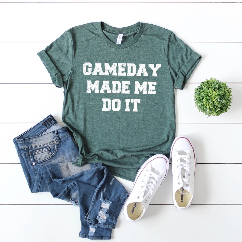 May include: A green t-shirt with the text "GAMEDAY MADE ME DO IT" in white distressed lettering. The shirt is paired with blue jeans and white sneakers.