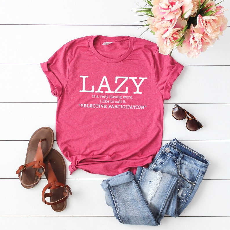 Op de afbeelding: Een roze T-shirt met de tekst "LAZY is a very strong word. I like to call it "SELECTIVE PARTICIPATION""