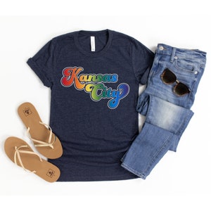 Pu&ograve; includere: Una maglietta blu navy con una grafica arcobaleno che dice "Kansas City" in un font retr&ograve;.