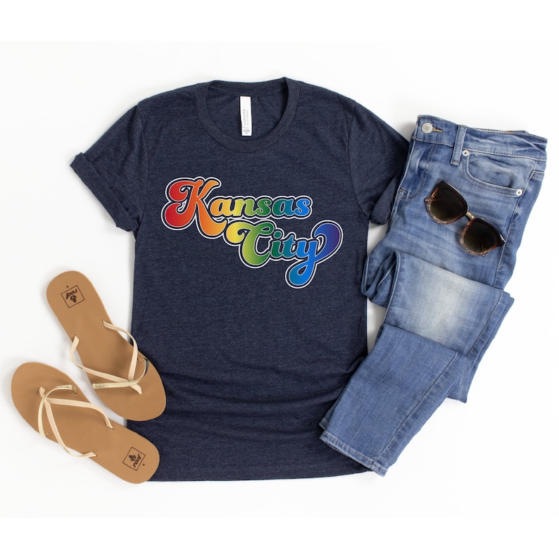 Pu&ograve; includere: Una maglietta blu navy con una grafica arcobaleno che dice "Kansas City" in un font retr&ograve;.