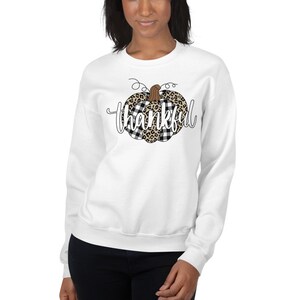 May include: White sweatshirt featuring a black and white checked and leopard print pumpkin design with the word "thankful" in white text.