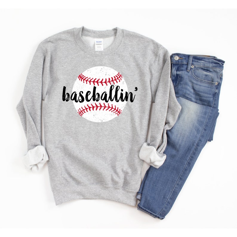May include: A light gray sweatshirt with a distressed baseball graphic and the word "baseballin'" in black script. The baseball is white with red stitching. Blue jeans are folded next to the sweatshirt.