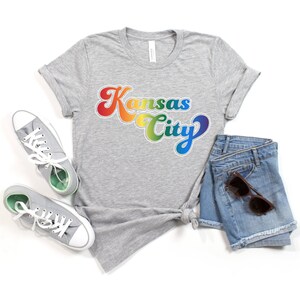 Pu&ograve; includere: Una maglietta grigia con una grafica arcobaleno che dice "Kansas City" in uno stile di font retr&ograve;. La maglietta &egrave; abbinata a pantaloncini di jeans e scarpe da ginnastica bianche.