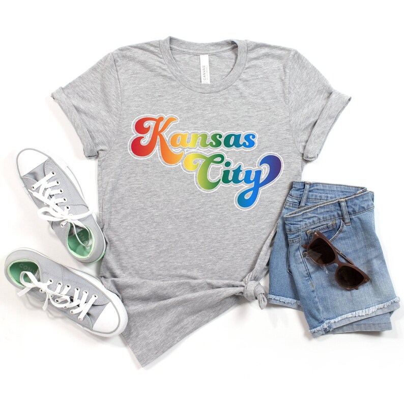Pu&ograve; includere: Una maglietta grigia con una grafica arcobaleno che dice "Kansas City" in uno stile di font retr&ograve;. La maglietta &egrave; abbinata a pantaloncini di jeans e scarpe da ginnastica bianche.