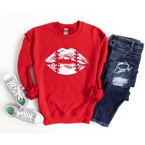 May include: Red sweatshirt with a white baseball-themed lips graphic. The sweatshirt is laid flat on a white background with a pair of gray sneakers and blue jeans.