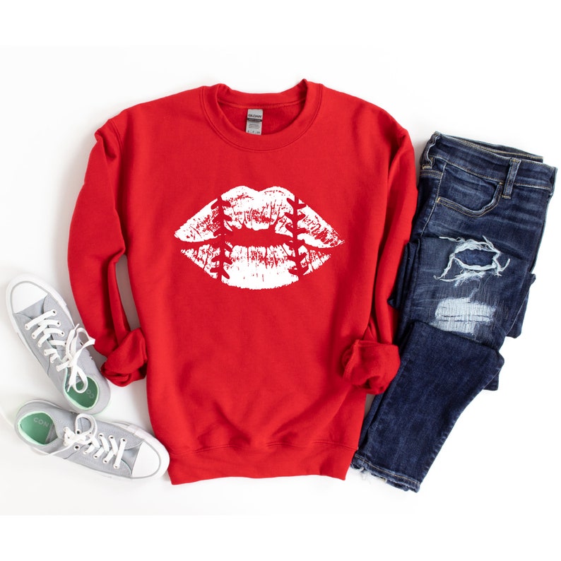 May include: Red sweatshirt with a white baseball-themed lips graphic. The sweatshirt is laid flat on a white background with a pair of gray sneakers and blue jeans.