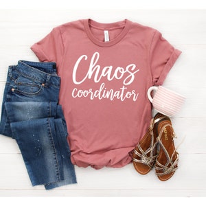 以下が含まれることがあります： 白い文字で「Chaos coordinator」と書かれた、くすんだピンクのTシャツ。白い木製の表面に、茶色のサンダル、青いジーンズ、ピンクと白のストライプのマグカップと一緒に置かれています。