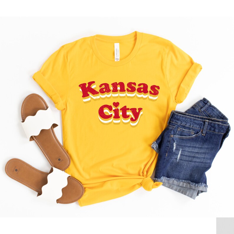 Pode incluir: Uma camiseta amarela com o texto "Kansas City" em letras retr&ocirc; vermelhas e brancas. A camiseta est&aacute; colocada sobre um fundo branco com um par de sand&aacute;lias brancas e shorts jeans azuis.