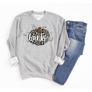 May include: A grey sweatshirt featuring a black and white checked and leopard print pumpkin design with the word "thankful" in white text.