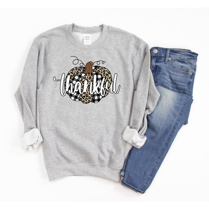 May include: A grey sweatshirt featuring a black and white checked and leopard print pumpkin design with the word "thankful" in white text.