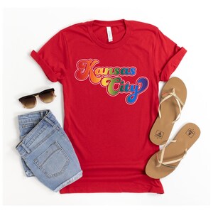 Pu&ograve; includere: Maglietta rossa con una grafica "Kansas City" arcobaleno. La maglietta &egrave; stesa su una superficie bianca con un paio di pantaloncini di jeans e un paio di sandali marroni.