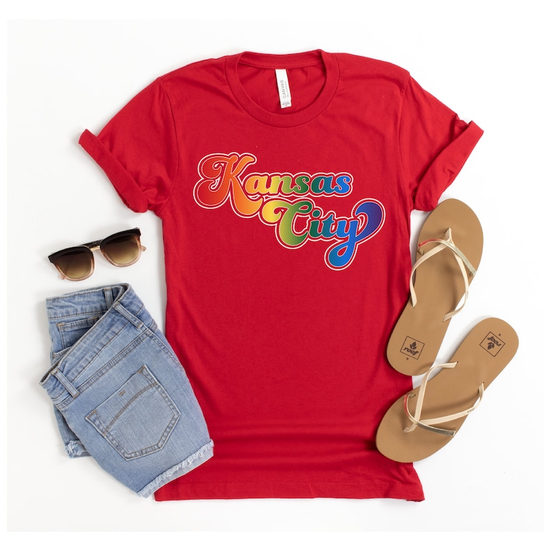 Pu&ograve; includere: Maglietta rossa con una grafica "Kansas City" arcobaleno. La maglietta &egrave; stesa su una superficie bianca con un paio di pantaloncini di jeans e un paio di sandali marroni.