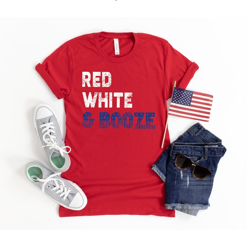 May include: Red, white, and blue graphic tee shirt with the text "Red White & Booze" printed on it. The shirt is red and the text is in blue and white. The shirt is laid flat on a white surface with a pair of grey sneakers, a pair of blue denim shorts, a pair of sunglasses, and a small American flag.