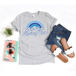 May include: Gray t-shirt with a blue and white rainbow graphic and the text "Mile High Anxiety Club" in a retro font.