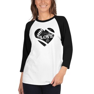 Może przedstawiać: Biało-czarna koszulka bejsbolowa z rękawami raglan z czarnym, przetartym nadrukiem w kształcie serca piłki nożnej z napisem "LOVE" w środku.