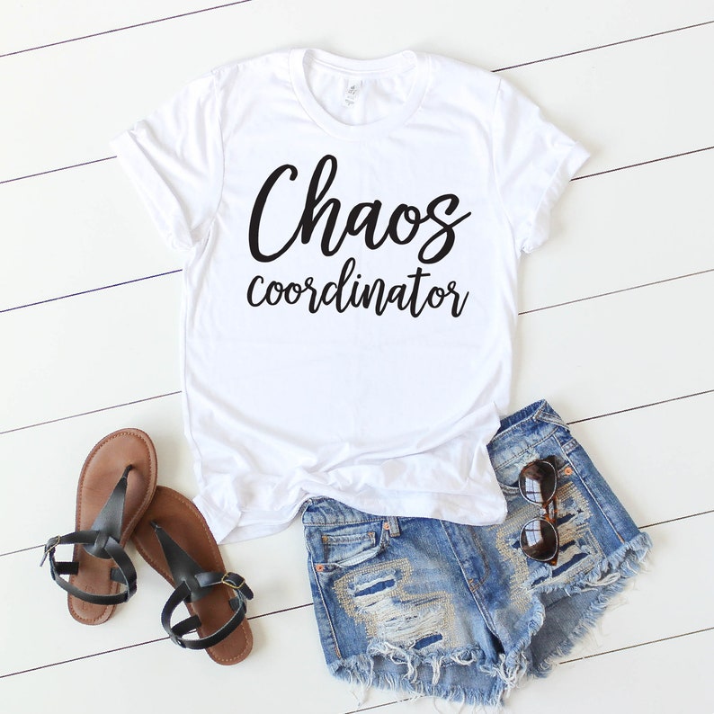 以下が含まれることがあります： 白いTシャツに黒い文字で「Chaos coordinator」と書かれています。Tシャツは白い木製の表面に置かれ、茶色のサンダルとデニムのショートパンツが置かれています。
