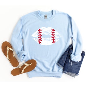 May include: A light blue sweatshirt with a white baseball-stitched lip design. The sweatshirt is folded and laid on a white surface with a pair of brown sandals and a pair of blue denim shorts.