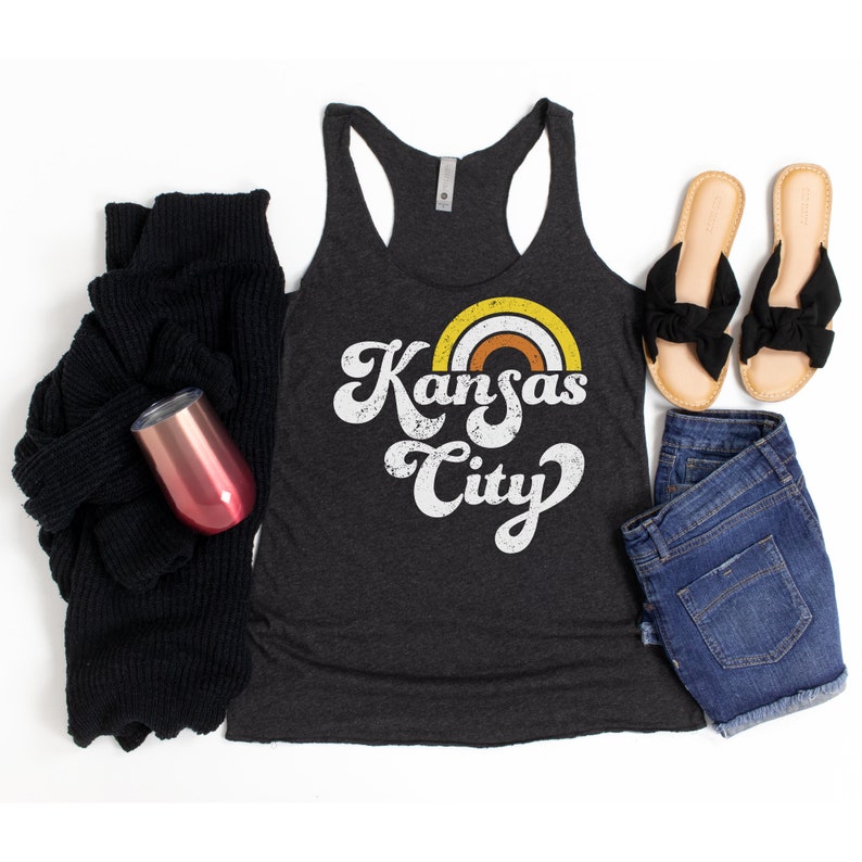 Puede incluir: Un top de tirantes gris oscuro con un gr&aacute;fico de estilo retro que dice "Kansas City" con un arco&iacute;ris sobre el texto. El top est&aacute; colocado sobre un fondo blanco con un su&eacute;ter negro, un vaso rosa, pantalones cortos de mezclilla azul y un par de sandalias marrones.