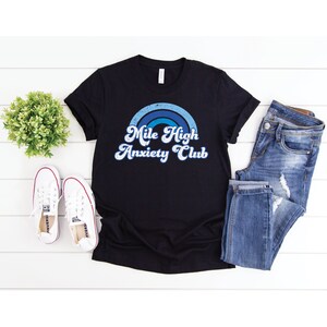 May include: Black t-shirt with a blue and white rainbow graphic and the text "Mile High Anxiety Club" in a retro font.