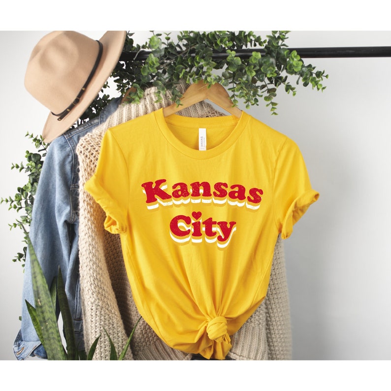 Pode incluir: Uma camiseta amarela com as palavras "Kansas City" em letras retr&ocirc; vermelhas e brancas.