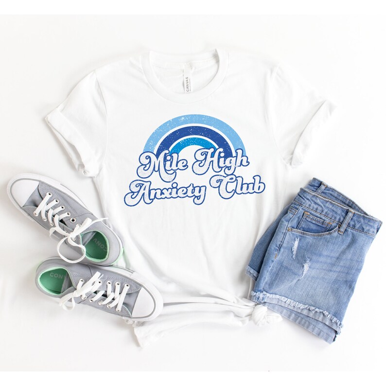 May include: White t-shirt with a blue and white rainbow graphic and the text "Mile High Anxiety Club" in a retro font.