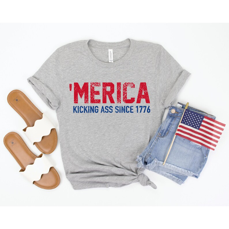 May include: A gray t-shirt with the text "'MERICA KICKING ASS SINCE 1776" in red and blue lettering. The shirt is laid out on a white surface with a pair of white sandals and a pair of denim shorts with a small American flag.