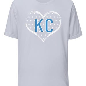Pode incluir: Camiseta azul claro com um design de cora&ccedil;&atilde;o branco com bolinhas azuis e as letras "KC" no centro.