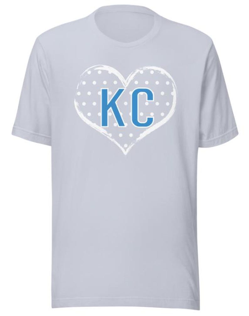 Pode incluir: Camiseta azul claro com um design de cora&ccedil;&atilde;o branco com bolinhas azuis e as letras "KC" no centro.