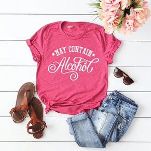 May include: A pink t-shirt with the text "*MAY CONTAIN* Alcohol" in white cursive lettering. The shirt is laid out on a white wooden surface with a pair of brown sandals, blue jeans, and sunglasses.