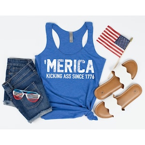 May include: A blue tank top with the text "'MERICA KICKING ASS SINCE 1776" in white. The tank top is laying flat on a white surface with a pair of blue denim shorts, red, white, and blue sunglasses, and a pair of brown sandals.