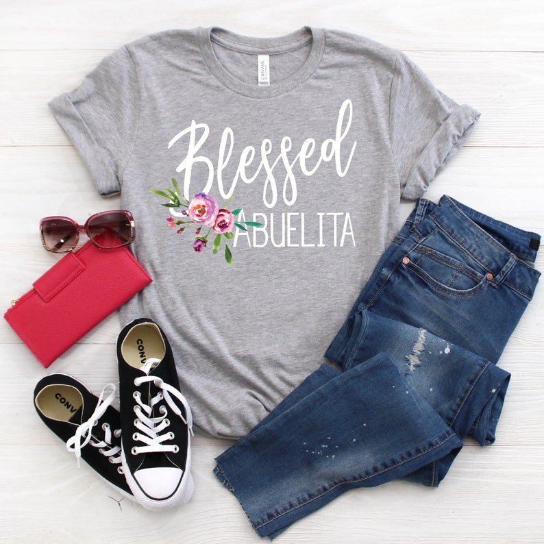 May include: Gray t-shirt with the text "Blessed Abuelita" in white lettering with a floral design in pink, purple, and green.