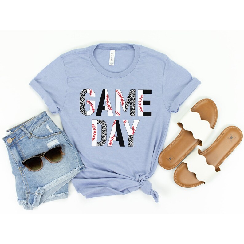 May include: A light blue t-shirt with the words "GAME DAY" printed on it. The letters are made up of a baseball pattern with red stitching.