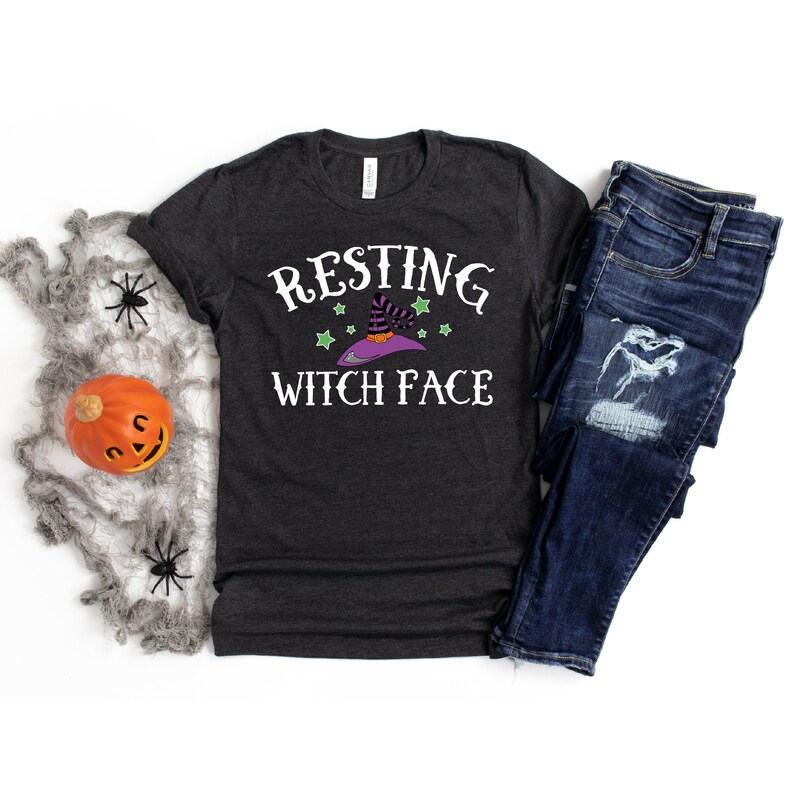 Puede incluir: Una camiseta gris oscuro jaspeada con el texto "Resting Witch Face" en blanco. Una bruja de dibujos animados con un sombrero morado y estrellas verdes est&aacute; por encima del texto.