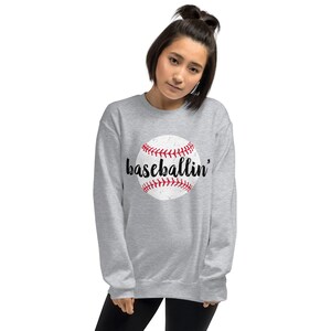 May include: A light gray sweatshirt featuring a distressed baseball graphic with red stitching and the word "baseballin'" in black script. The sweatshirt has a crew neck and long sleeves.