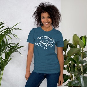 May include: A woman wearing a teal blue t-shirt with the text "*MAY CONTAIN* Alcohol" in white cursive lettering.