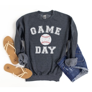 May include: A dark grey sweatshirt with the text "GAME DAY" in white with a vintage baseball graphic in the centre. The sweatshirt is laid out on a white background with a pair of brown sandals and a pair of blue denim shorts.