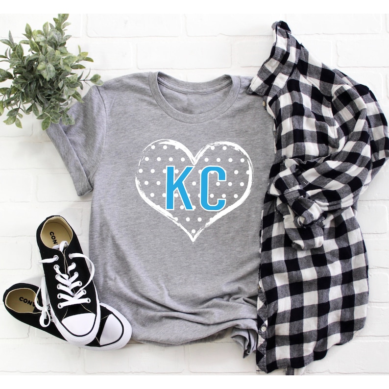 Pode incluir: Camiseta cinza com um design de cora&ccedil;&atilde;o branco com bolinhas azuis e as letras "KC" em azul.