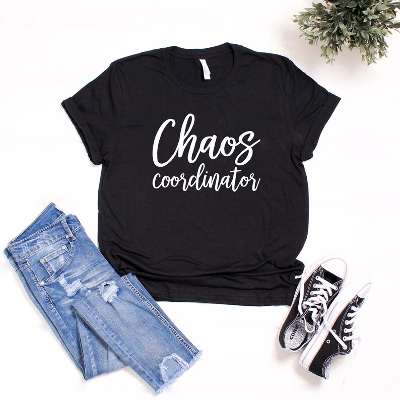 以下が含まれることがあります： 白い文字で「Chaos coordinator」と書かれた黒いTシャツ。白い背景に、青いジーンズと黒いスニーカーと一緒に置かれています。
