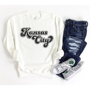 May include: White sweatshirt with a black retro-style graphic that says "Kansas City". The sweatshirt is shown with a pair of blue jeans and gray sneakers.