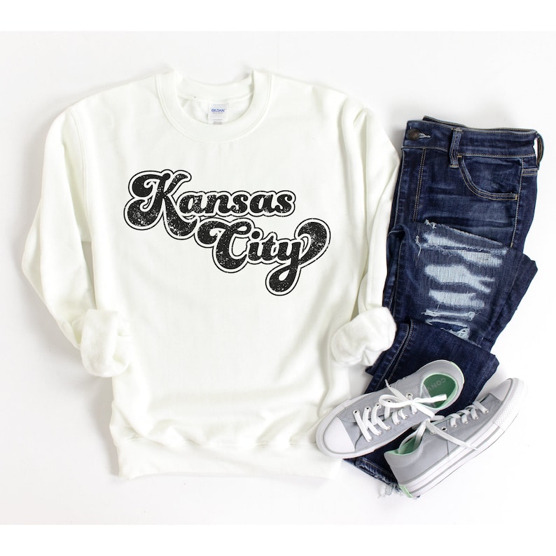 May include: White sweatshirt with a black retro-style graphic that says "Kansas City". The sweatshirt is shown with a pair of blue jeans and gray sneakers.