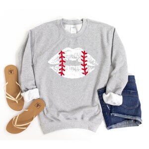 May include: A gray sweatshirt with a white baseball-stitched lips design. The sweatshirt is shown with a pair of brown sandals and denim shorts.