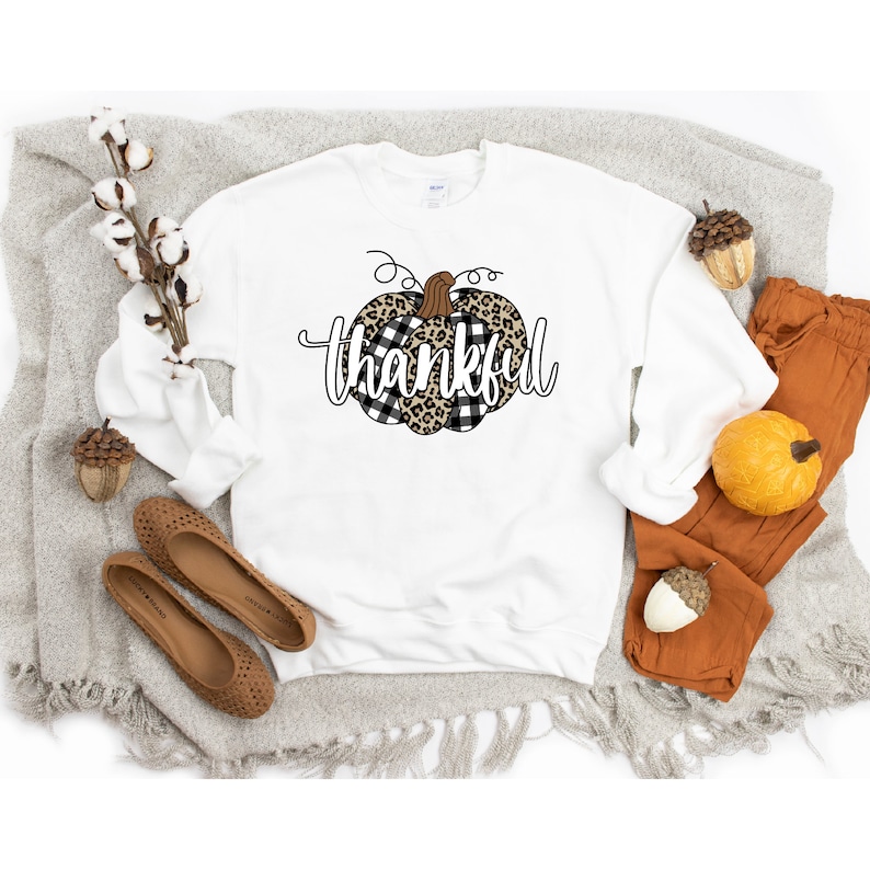 May include: White sweatshirt featuring a black and white checked and leopard print pumpkin design with the word "thankful" in black script.