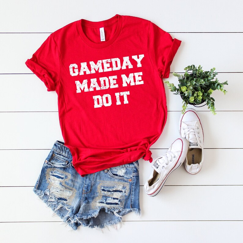 May include: Red t-shirt with white text that reads "GAMEDAY MADE ME DO IT". The shirt is paired with denim shorts and white sneakers.