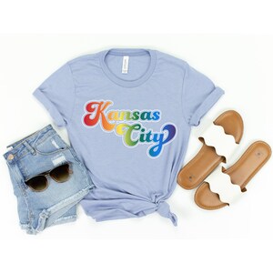 Pu&ograve; includere: Una maglietta blu chiaro con una grafica arcobaleno che dice "Kansas City" in uno stile di font retr&ograve;. La maglietta &egrave; mostrata su uno sfondo bianco con un paio di sandali bianchi e pantaloncini di jeans.
