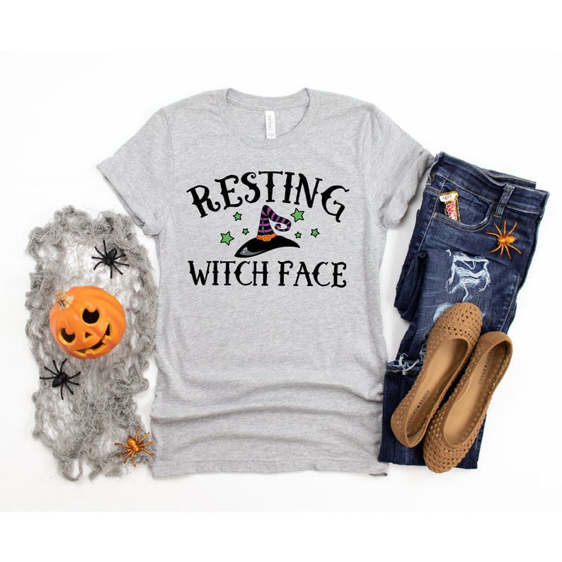 Puede incluir: Una camiseta gris con el texto "Resting Witch Face" en negro. Un sombrero de bruja con estrellas verdes est&aacute; por encima del texto. La camiseta est&aacute; colocada sobre un fondo blanco con un par de vaqueros y zapatos marrones.