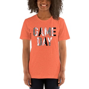 May include: A bright orange t-shirt with the words "GAME DAY" printed in black letters. The letters are partially covered with a black and white baseball pattern.