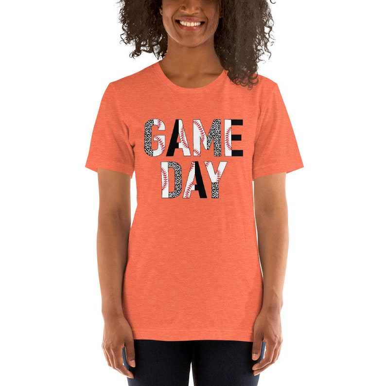 May include: A bright orange t-shirt with the words "GAME DAY" printed in black letters. The letters are partially covered with a black and white baseball pattern.